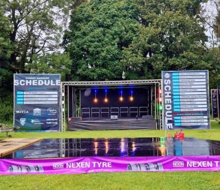 custom build stage for car show Stage setup with lighting and event schedule posters in an outdoor setting.